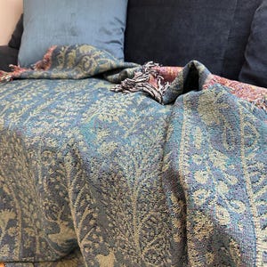 May include: A woven throw blanket featuring a detailed forest scene with deer and trees in shades of teal, blue, and gold. The fabric has a textured appearance with fringed edges, displaying a repeating pattern of woodland animals and foliage.