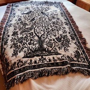 May include: A rectangular woven throw blanket featuring a black and cream botanical design of a large tree with birds perched on its branches. The blanket has a decorative border and frayed edges along the top and bottom sides of the fabric.