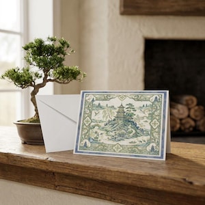 Puede incluir: Una tarjeta de felicitación rectangular con una ilustración detallada de una pagoda en una isla rocosa rodeada de agua, árboles y aves, enmarcada por un borde decorativo en tonos azules y verdes, acompañada de un sobre blanco liso.
