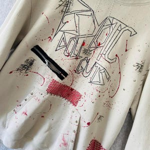 May include: Cream-coloured hoodie with black and red graphic design elements. Features a black zip, red mesh accents, and silver studs. The text "ALL OUT GUNS" is prominently displayed in black.