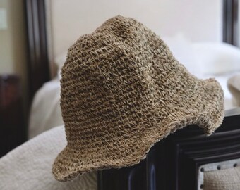 May include: A tan bucket hat crafted from a textured, woven material. The hat features a rounded crown and a short, slightly downturned brim. The construction displays a consistent, open-knit pattern throughout the entire surface of the headwear.
