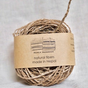 May include: A ball of natural fiber twine with a brown paper label wrapped around it. The label reads "HUMLA HANDMADE natural fibers made in Nepal". The twine is a light brown color and appears to be made of jute or a similar material.