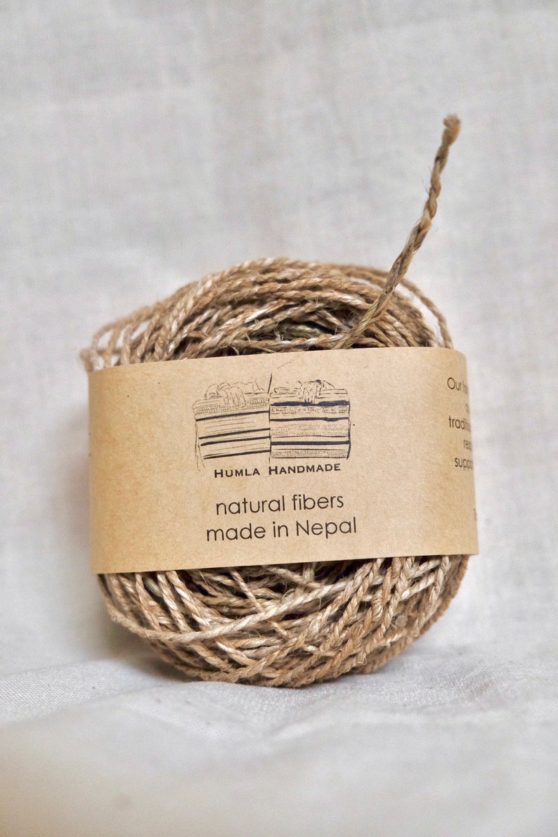 May include: A ball of natural fiber twine with a brown paper label wrapped around it. The label reads "HUMLA HANDMADE natural fibers made in Nepal". The twine is a light brown color and appears to be made of jute or a similar material.