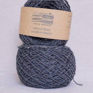 May include: Two gray yarn balls stacked on top of each other. The top ball has a brown label with the text "HUMLA HANDMADE natural fibers made in Nepal". The yarn appears to be made of natural fibers.