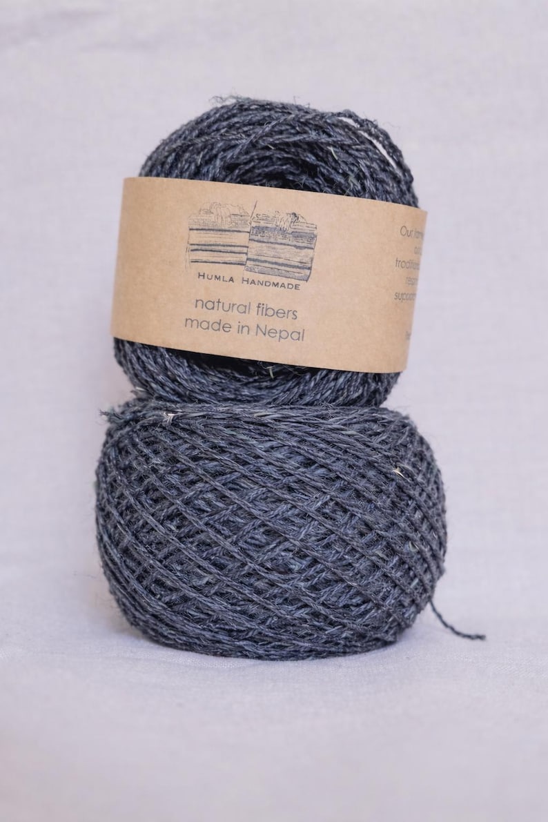 May include: Two gray yarn balls stacked on top of each other. The top ball has a brown label with the text "HUMLA HANDMADE natural fibers made in Nepal". The yarn appears to be made of natural fibers.