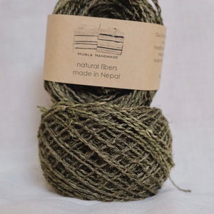 May include: Two spools of natural fiber yarn in shades of green and brown, with a label that reads "HUMLA HANDMADE natural fibers made in Nepal". The yarn is tightly wound, showing the texture of the fibers.