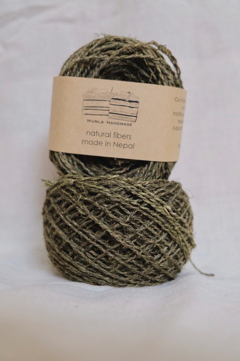 May include: Two spools of natural fiber yarn in shades of green and brown, with a label that reads "HUMLA HANDMADE natural fibers made in Nepal". The yarn is tightly wound, showing the texture of the fibers.