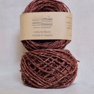 May include: Two balls of brown natural fiber yarn, one atop the other. A label on the top ball reads "HUMLA HANDMADE natural fibers made in Nepal". The yarn has a textured appearance, suitable for knitting or other fiber crafts.
