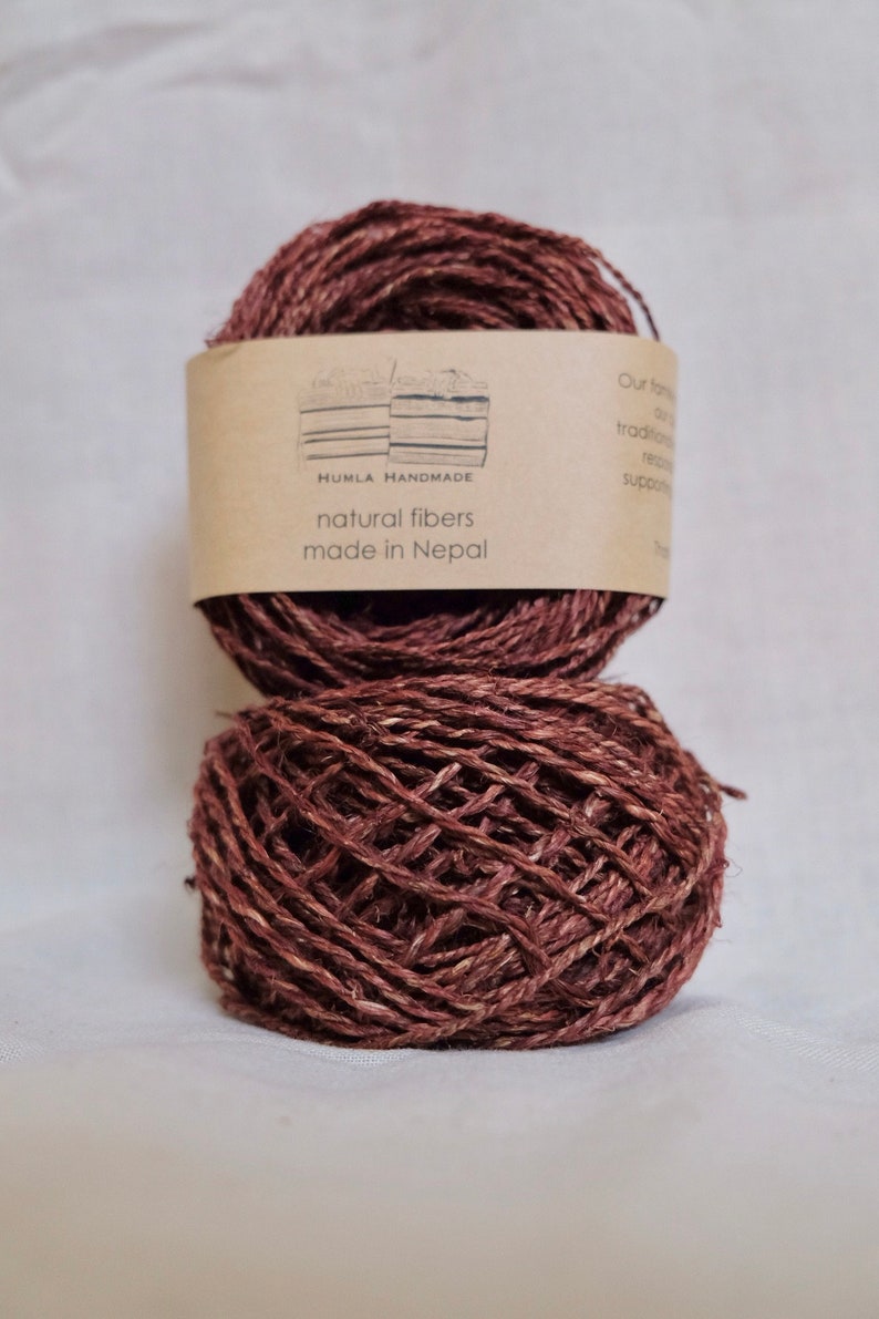 May include: Two balls of brown natural fiber yarn, one atop the other. A label on the top ball reads "HUMLA HANDMADE natural fibers made in Nepal". The yarn has a textured appearance, suitable for knitting or other fiber crafts.