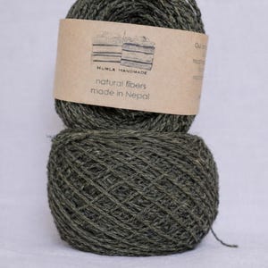 May include: Two olive green yarn skeins are stacked on a white background. A brown label on the top skein reads "HUMLA HANDMADE natural fibers made in Nepal". The yarn appears to be a natural fiber, possibly wool or cotton.