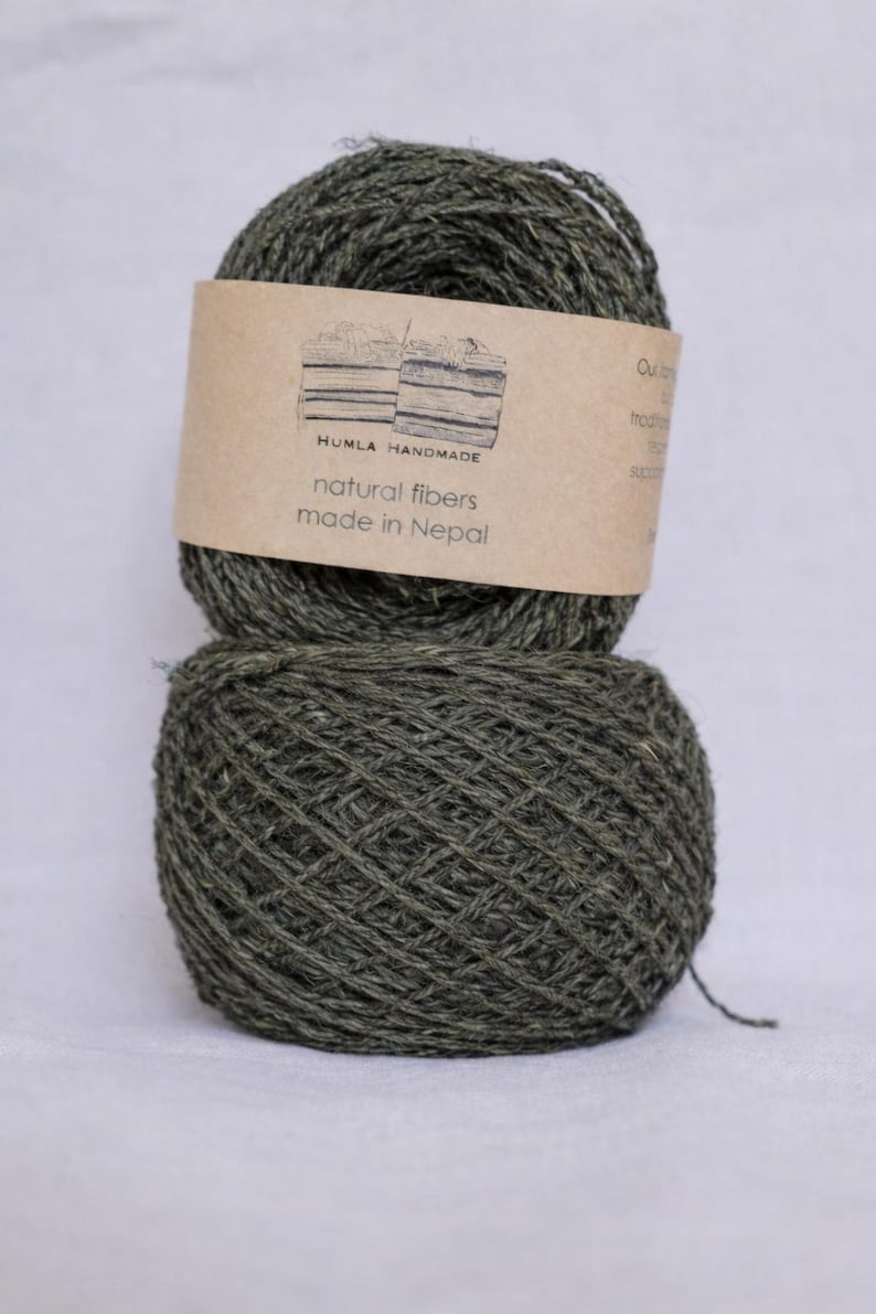 May include: Two olive green yarn skeins are stacked on a white background. A brown label on the top skein reads "HUMLA HANDMADE natural fibers made in Nepal". The yarn appears to be a natural fiber, possibly wool or cotton.