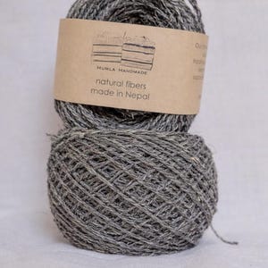 May include: Two gray yarn balls are stacked on top of each other. A brown label is wrapped around the top ball, with the text "HUMLA HANDMADE natural fibers made in Nepal". The yarn appears to be made of natural fibers.