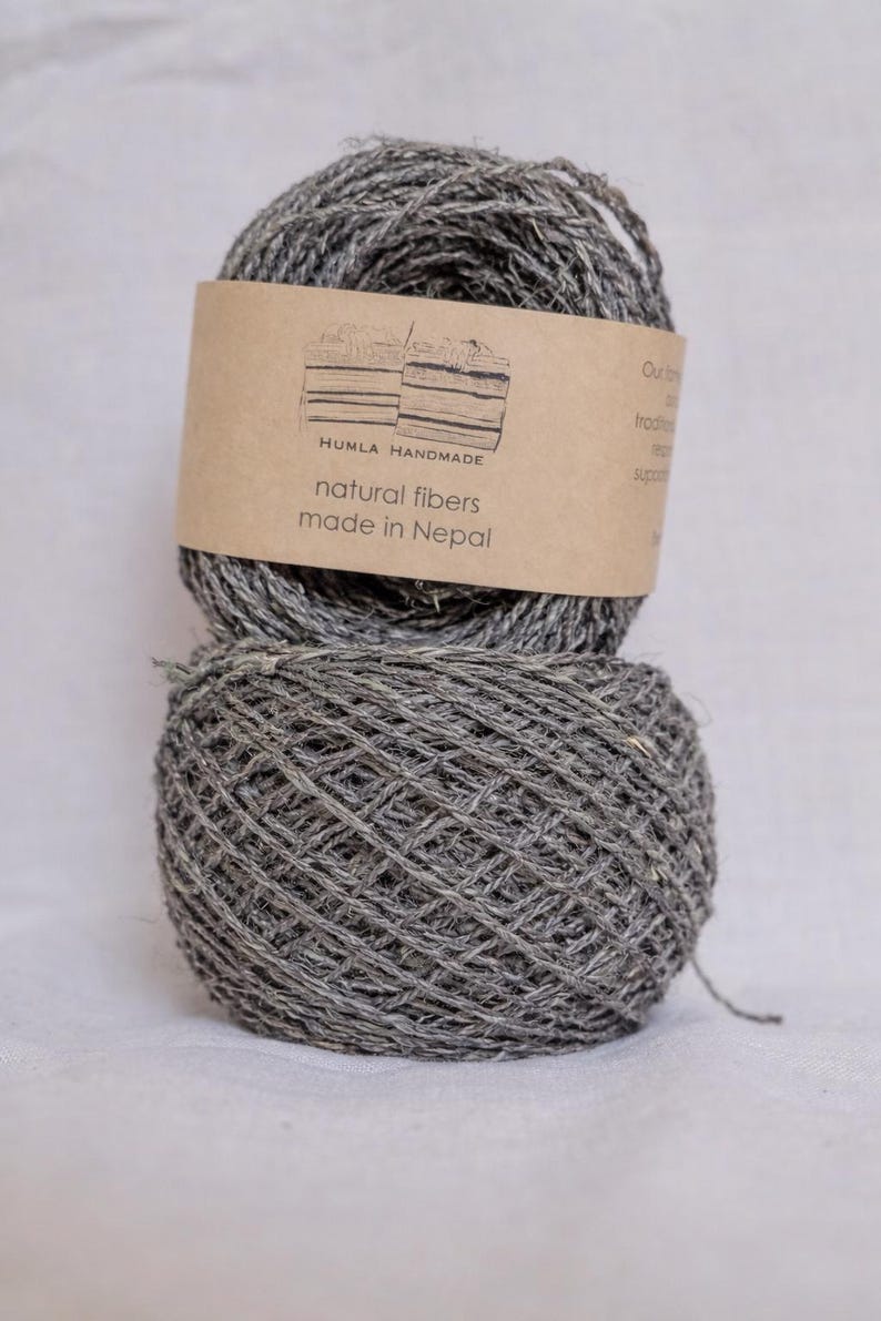 May include: Two gray yarn balls are stacked on top of each other. A brown label is wrapped around the top ball, with the text "HUMLA HANDMADE natural fibers made in Nepal". The yarn appears to be made of natural fibers.