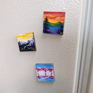 May include: Three small, square magnets with miniature landscape paintings. One depicts mountains under a yellow sky, another a sunset over a lake, and the third features pink trees reflected in water. Each is painted with visible brushstrokes.