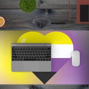 Op de afbeelding: Een bureaumat met een non-binaire pride vlag ontwerp, een laptop, muis en koffiekopje. De mat heeft een geel, wit, paars en zwart kleurenschema. Een notitieboek, pen en kleine plant zijn ook zichtbaar.