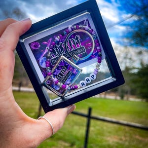 Puede incluir: Un marco negro con un inserto de plástico transparente que contiene un dije morado y plateado con el texto "The Real Project" y una pulsera de cuentas moradas y plateadas con el texto "RRP".