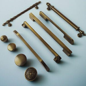 May include: A selection of antique brass cabinet hardware, including handles and knobs. The handles have different designs, some with textured or ornate details. The knobs are round and feature various patterns.
