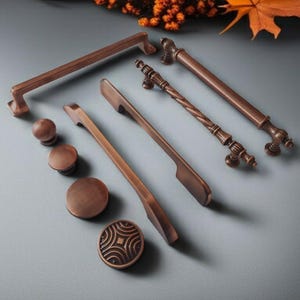 Op de afbeelding: Een verzameling bronzen kastbeslag, waaronder handgrepen en knoppen. De set bevat verschillende ontwerpen, van eenvoudige stangtrekkers tot sierlijke, gedetailleerde handgrepen. De hardware is gerangschikt op een grijze ondergrond, met herfstbladeren op de achtergrond.