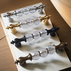 May include: Four cabinet pulls with clear crystal-like center sections and decorative metal ends. The pulls have silver, gold, black, and antique brass finishes. They are arranged on a white surface, showcasing their design and color variations.