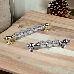 Op de afbeelding: Twee kristallen kastknoppen met een facetgeslepen ontwerp. De ene knop is goud en de andere is zilver.