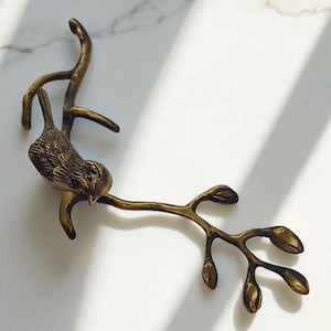 May include: A bronze-coloured decorative bird perched on a branch with leaves. The bird has detailed feathering and is attached to a curved branch with several small leaves. The piece is set against a white marble background.