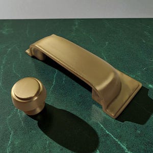 Puede incluir: Un juego de herrajes dorados para armarios, que incluye un pomo y un tirador. El pomo es redondo con la parte superior plana, y el tirador es rectangular con un asa curvada. Los herrajes se muestran sobre una superficie verde marmolada.