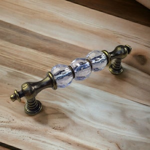 以下が含まれることがあります： 透明なガラスのデザインの真鍮製のキャビネットプル。ガラスはファセットカットされており、薄い青色をしています。プルは木製の表面に取り付けられています。