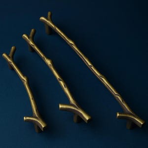Op de afbeelding: Drie bronskleurige kastgrepen in een takontwerp, tegen een donkerblauwe achtergrond. De grepen variëren in lengte, de langste meet ongeveer 25 cm. Het getextureerde oppervlak bootst het uiterlijk van natuurlijke takken na.
