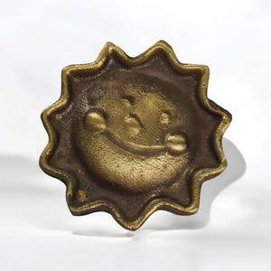 Peut inclure: Un objet décoratif en forme de soleil de couleur bronze avec un visage souriant. Le soleil a un motif en forme d'éclat d'étoile autour du visage. L'objet est probablement en métal et a un aspect vintage ou antique.