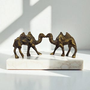May include: Two bronze camel figurines facing each other, positioned on a white marble rectangular base. The camels are detailed with textured surfaces and stand in a pose that suggests interaction. The background is a plain white wall.