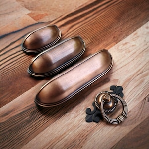 Op de afbeelding: Drie antieke bronzen kastgrepen en een enkele bronzen ringgreep met een zwarte metalen basis. De grepen zijn ovaalvormig met een licht verhoogde rand. De ringgreep heeft een decoratief krulontwerp.