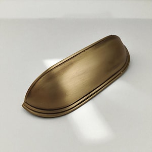 Puede incluir: Un tirador de armario dorado en forma de copa. El herraje tiene una forma curvada con tres líneas paralelas cerca del borde inferior. El acabado tiene un aspecto ligeramente envejecido.