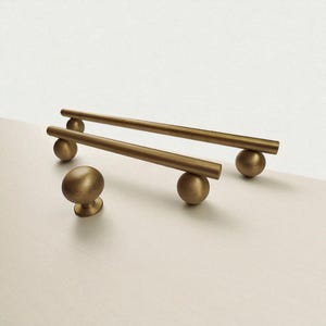 May include: A set of gold-toned cabinet hardware, including two bar pulls and a round knob. The bar pulls are supported by spherical bases, and the knob has a rounded top and a small, circular base. The hardware is displayed on a light-colored surface.