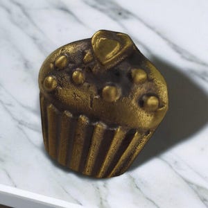 Könnte beinhalten: Ein bronzefarbenes, cupcake-förmiges Dekorationsobjekt. Der Cupcake hat ein herzförmiges Detail oben und kleine, runde Verzierungen. Das Objekt befindet sich vor einem weißen Marmorhintergrund.