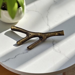 May include: A bronze-colored decorative branch with three prongs, resting on a white marble-patterned surface. The branch has a textured, naturalistic appearance. A small white pot with a green plant is in the background.