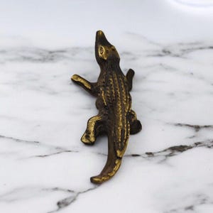 May include: A detailed bronze alligator figurine, with textured skin and outstretched limbs. The sculpture is positioned on a white and gray marble surface, showcasing the intricate details of the reptile.