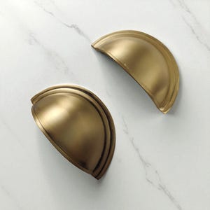 Może przedstawiać: Dwa uchwyty do szafek w kształcie filiżanki w kolorze antycznego mosiądzu. Uchwyty mają kształt półksiężyca z zaokrąglonym wzorem. Wykończenie ma lekko postarzany wygląd, sugerujący styl vintage lub retro. Odpowiednie do szafek kuchennych lub łazienkowych.