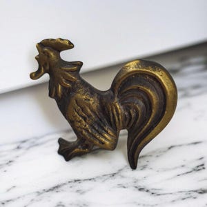 Könnte beinhalten: Eine bronzefarbene Hahnfigur mit detaillierten Federn und Kamm. Die Skulptur steht auf einer weiß-grauen Marmoroberfläche, die ihren antiken Charakter hervorhebt. Der Hahn ist ein dekoratives Objekt.