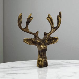 May include: A bronze-coloured deer head sculpture with detailed antlers and facial features. The sculpture has a textured surface and is set against a neutral background. The deer head is a decorative item.