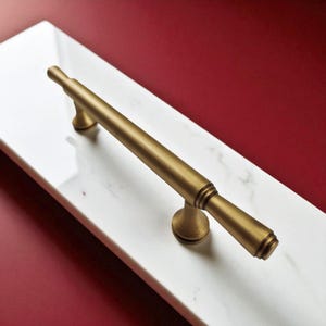 以下が含まれることがあります： 白い大理石の表面に置かれた、金色のキャビネットプルハンドル。 ハンドルは、装飾的なエンドキャップが付いた円筒形のバーで、2つの円形のベースに取り付けられています。 背景は無地の赤色です。