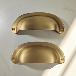 May include: Two antique brass-coloured cup pull handles. Each handle has a semi-circular design with two screw holes on each end. The handles are made of metal and have a slightly aged appearance, suggesting a vintage or rustic style.