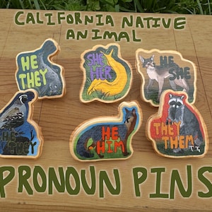 May include: Wooden pronoun pins featuring California native animals. Each pin displays a different animal with corresponding pronouns: He/They, She/Her, He/She, and They/Them. The pins are set on a wooden surface with the text "CALIFORNIA NATIVE ANIMAL" and "PRONOUN PINS".