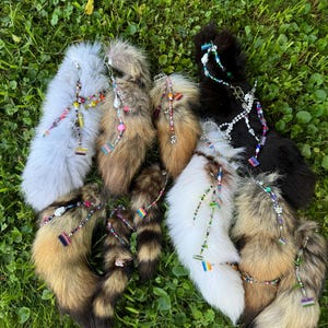 May include: A selection of faux fur tails in shades of grey, brown, white, and black, decorated with beaded details and rainbow pride flag charms. The tails are displayed on a bed of green grass.