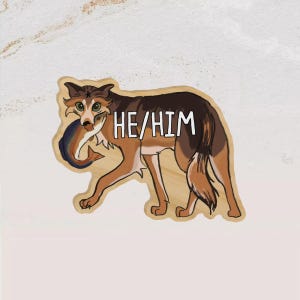 May include: A die-cut sticker featuring a brown, black, and tan canine illustration holding a fish in its mouth. The text HE/HIM is printed in white capital letters across the body of the canine. The sticker has a light wood-grain texture background.