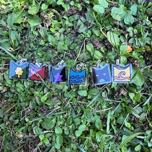 May include: Six clear acrylic charms with colourful designs, such as a tree, lightning bolts, and waves. Each rectangular charm has a silver metal loop and a black cord. The charms are displayed on green grass. The designs are in various colours.
