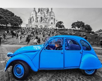 Op de afbeelding: Een felblauwe vintage auto rijdt door een zwart-witte Parijse straat. De auto is scherp, terwijl de achtergrond wazig is. De auto rijdt langs een grote kerk.