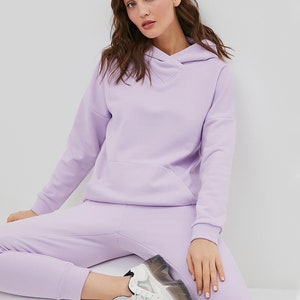 May include: A light purple hooded sweatshirt with long sleeves, a front kangaroo pocket, and a crossover neckline, paired with matching light purple sweatpants and white sneakers with a thick, translucent, and black-accented rubber sole.