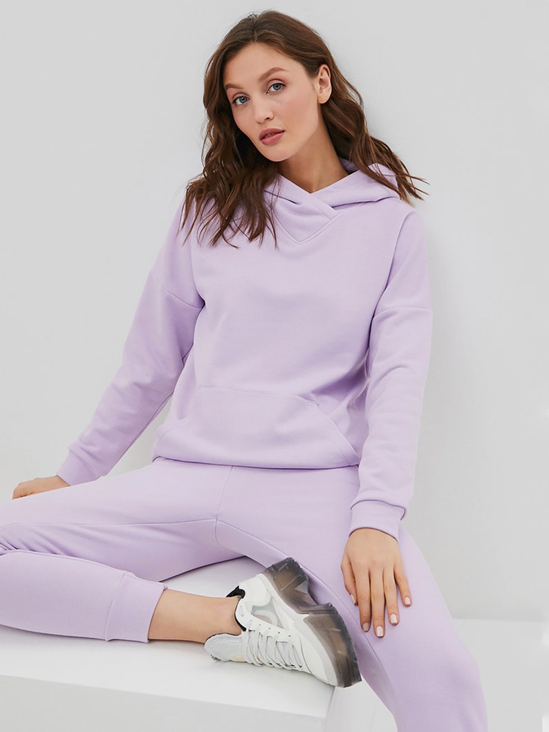 May include: A light purple hooded sweatshirt with long sleeves, a front kangaroo pocket, and a crossover neckline, paired with matching light purple sweatpants and white sneakers with a thick, translucent, and black-accented rubber sole.