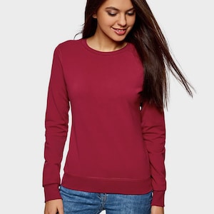 May include: A solid burgundy long-sleeve sweatshirt with a crew neckline and ribbed cuffs, paired with blue denim jeans. The sweatshirt features a simple, plain design with a fitted hemline that sits at the waist, providing a clean look.