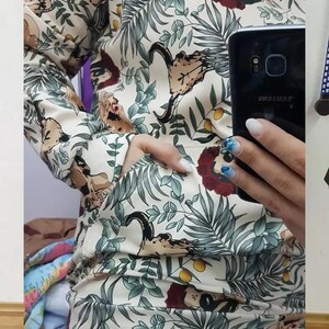 May include: A long-sleeve hoodie in an off-white color featuring a repeating pattern of green tropical leaves, yellow citrus fruits, and stylized skulls adorned with red floral crowns. The garment includes a front kangaroo pocket and ribbed cuffs.